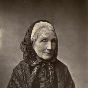 Black and white, head-and-shoulders photograph of the aged Mary Howitt. She is dressed in black: a dark hood covers her grey hair and white cap; her black, patterned cloak, tied with dark ribbons, covers a dark dress buttoned in front. Her eyes are cast down.