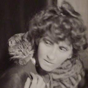 Black and white, head-and-shoulders photograph of Violet Hunt with a tabby cat perched on her shoulder. She has short, curly hair, and is wearing a simple, dark shirt.