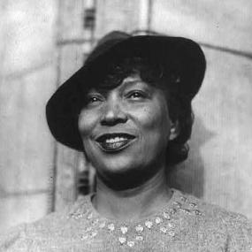 Black and white, head-and-shoulders photograph of Zora Neale Hurston smiling. She stands, wearing a small  black hat and a sweater with decorated neckline. Library of Congress.