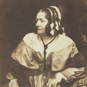 Sepia-toned photograph of Anna Brownell Jameson, standing with her elbow resting on a book that sits atop a small cloth-draped table. She wears a light dress with white lace trim around the neck, a second layer over the shoulders, apparently a built-in shawl trimmed with dark gauzy cloth. A long, dark, gauzy scarf is draped behind her and over her arms. Her dark curly hair is topped by a bonnet of light and dark ribbons. In one hand she holds a small pair of spectacles.