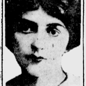 Black and white photograph of F. Tennyson Jesse, shown from the shoulders up, in a shape with symmetrically ornamental corners. The picture, with strong light and dark contrast, shows her wearing a small hat or headband on short, dark, bushy hair parted in the middle, and a white blouse with black bow.