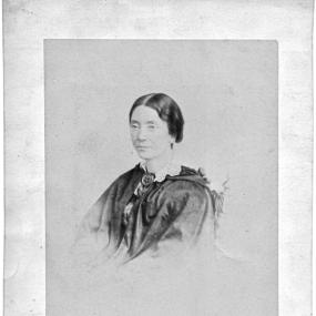 Head-and-shoulders photograph of Geraldine Jewsbury. She is wearing a dark, hooded cloak, with a light lace collar and a rose-shaped fastener. Her dark wavy hair is pulled back. Her full signature is reproduced underneath: "Geraldine Endsor Jewsbury".