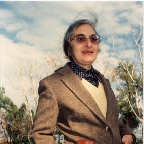 Colour photo of Ruth Prawer Jhabvala at the home of her frequent collaborator, director James Ivory, in Claverack, New York, September 1981.
            She is wearring a tweed blazer and has her hands in her pockets. She is standing in a park in fall, and the sky is blue with many clouds. The are birch trees in the background are
            bare.