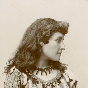 Black and white photograph of Pauline Johnson, shown from the waist up. She wears a necklace of bear claws and a loose shirt with leather fringes and ermine tails. Her dark hair is curled in front and hangs loose down her back.