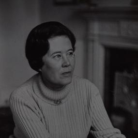 Black and white photograph of Pamela Hansford Johnson, seated, with her hands clasped, wearing a white polo-neck sweater with a brooch on the collar. Her dark, wavy hair is smooth and short.