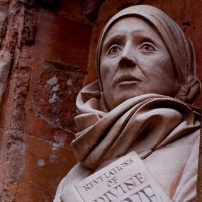 Photograph of the statue of Julian of Norwich sculpted for a niche in the west front of Norwich Cathedral by David Holgate in 2014, six centuries after Julian's death. The photo, taken from below, shows only the waist up. Julian has an alert face and heroic stance, wrapped in a loose cloak and hood, and holding a book titled (in capitals very unlike the writing of her own day) "Revelations of Divine Love."
