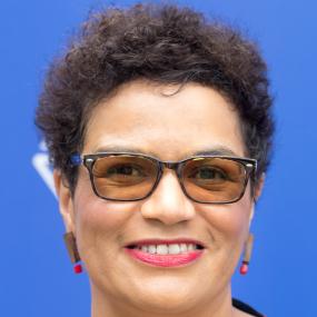 Photo of Jackie Kay taken during the annual Edinburgh International Book Festival. She is wearing brown tinted glasses and a dress with small
            polka-squares that matches her earrings. she has short black hair.