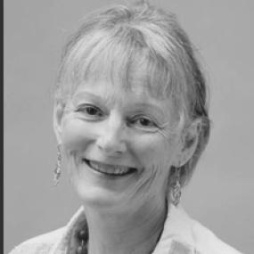Black and white, head-and-shoulders, smiling photograph of Judith Kazantzis. She wears a light collared shirt in a muted print a necklace of large beads, and simple dangling earrings. Her blonde hair has a long fringe.