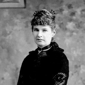 Black and white photograph of Anna Kingsford, standing in an interior setting with a frame on the wall and a chair behind her.  She is wearing a black lace dress with a black ribbon around her neck, black gloves, and a black clutch in her hands. Her light hair is twisted and pinned on top of her head, and she has short curly bangs.