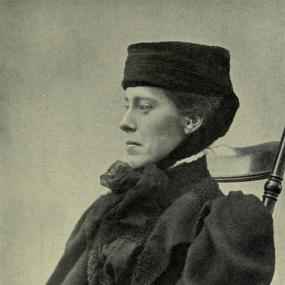 Black and white photograph of Mary Kingsley in profile, seated in a wooden chair. She is wearing a black pillbox hat that ties under her chin, and a dark dress with leg-of-mutton sleeves.