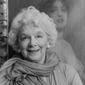 Black-and-white photo of Rosamond Lehmann. She is wearing a silk scarf, and her white hair is cut short. Behind her is a painting of a woman. 