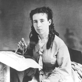 Black and white photograph of Alice Dixon Le Plongeon, seated, holding a caliper compass against a large piece of paper or fabric. She is wearing a long dress, decorated with buttons and ruffles, and tied with a bow at the neckline. Her hair is partially pulled back and partially left down in dark curls.