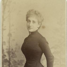 Black and white photograph of Ada Leverson, standing with her hands behind her back. She is wearing a dark dress with a high collar, long sleeves, a broad skirt, and a row of buttons up the front. Her hair is short and curly. Below her photograph are the words: "Elliot and Fry, 55 and 56, Baker St London. W", typed in all capitals.