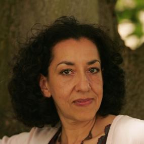 Portrait photo of Andrea Levy. She has curly black hair and a white cardigan. Behind her is a tree trunk and to her left is a smattering of
            foliage.