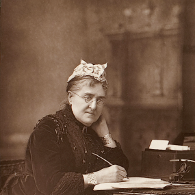 Photo of a carbon print of Eliza Lynn Linton by W. and D. Downey, published by Cassell, 1890. She sits at a small chinoiserie table with lattice-style legs, as if paused in the act of writing on a sheaf of papers. Her right hand holds a pen; her chin rests on her left. She has a broad face with wire spectacles perched on her nose, and wears a white cap and a dark (black?) dress in several layers, with lace at the wrists and a good deal of dark decoration including jet beads sewn on. She has a metal cuff bra