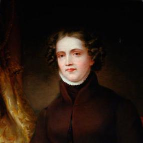 Photograph of a painting of Anne Lister by an unidentified artist; it hangs at Lister's estate, Shibden Hall. She is shown from the waist up against the background of a gold curtain, wearing a high-collared maroon jacket. Her jaw-length dark hair, smoothly parted in the middle, is curly around her face.