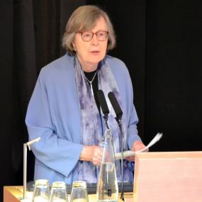 Colour photo, half-length, of Penelope Lively talking at a microphone, with a water carafe and tumblers in front. She is wearing tortoiseshell glasses, a blue jacket over black, a scarf, and a silver chain. Her grey hair is jaw-length.