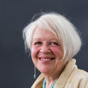 Portrait photo of Liz Lochhead. She is wearing a beige blazer and a turqoise beaded necklace. Her white hair is cut short and she is standing
            against a blue/grey backdrop.