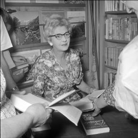 Black-and-white photo of Norah Lofts sitting at a desk for a book signing. Behind her are stacks of bookstore shelves. She has cat-eye glasses
            and wears a floral dress. People are gathered around her holding her books out to be signed. 