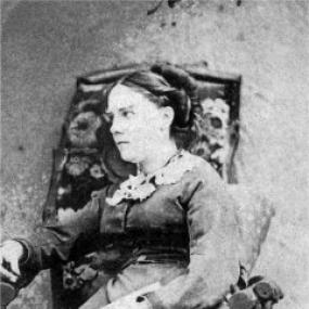 Black and white photograph of Maria Theresa Longworth, seated in a high-backed upholstered chair. She is wearing a simple, long, dark dress with a white lace collar, and her dark hair is pulled into a neat bun.
