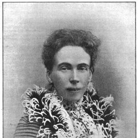 Black and white photograph of Edna Lyall by the firm of G. P. Abraham of Keswick. She is shown from the shoulders up, wearing a black and white feather boa over a dress which apparently has a white and black pattern on the body and stripes on the sleeves, She has dark, wavy hair.