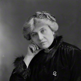 Black and white photograph of Edith Lyttelton, wearing a dark velvet dress with a fur trimmed collar and sleeves, with buttons on the shoulders. She is wearing a choker necklace with a black ribbon, and she has a bow in her light hair, which is pulled back. She has an elbow propped on a table and her head rests on that hand.
