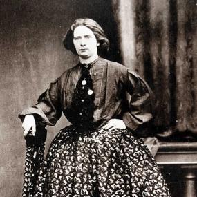 Black and white photograph of Jessie White Mario, standing with one hand on her hip and the other on the back of a carved chair. She is wearing a long skirt with a bold pattern and a simple, solid-coloured jacket with broad sleeves. Her hair is chin-length and her expression challenging.