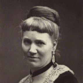 Black and white, head-and-shoulders photograph of Florence Marryat. She is wearing a dark velvet dress with a square lace collar. Her hair is worn in a bun; earrings dangle from her ears, and a necklace is tied tightly around her neck with a black ribbon. She holds a fan against her chest. Her name appears below in capitals in the form "Miss Florence Marryatt".