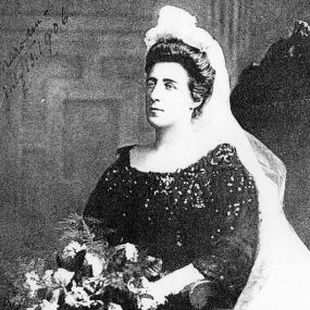 Black and white photo of Kate Marsden, 1906, shown from the waist up, wearing a dark dress speckled with ornament, with her dark hair pulled up and topped by a white headdress and long veil pushed back. She holds a bouquet. Her name is written in a top corner: "Marsden, July 13. 1906."