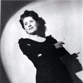 Black and white photo of Ngaio Marsh, standing in a spotlight and tilted backwards. Her black dress and dark hair are dramatically lit, and her hand holding a cigarette is doubled by its shadow behind.