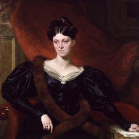 Photograph of the painting of Harriet Martineau by Richard Evans, 1833-4. She is seated on a small settee upholstered in red, with red drapery behind. She has one hand outstretched to rest on papers which sit atop a table with patterned tablecloth. She is wearing a low-necked long dress in dark blue satin, with huge puff sleeves and a gauzy white collar with a pearl pin in the centre, and a very long fur stole. Her shiny dark hair is centre-parted and piled on top of her head. National Portrait Gallery.