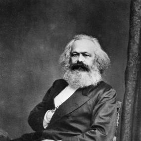 Black-and-white photograph of Karl Marx by John Jabez Edwin Mayal, before August 1875. Marx, with bushy hair, beard, and moustache, sits looking directly at the viewer, with his right hand tucked into the breast of his jacket.