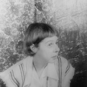 Black and white photograph of Carson McCullers, shown from the shoulders up, against a backdrop of flower-patterned drapery. She has jaw-length hair with short bangs, and she is wearing a baggy collared shirt with lines of decorative stitching up the front, boxy shoulders, and wide frayed sleeves.