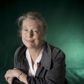 Portrait of Candia McWilliam. She is wearing a grey jacket and a white eyelet top and has a tartan pin at her lapel. 