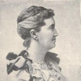 Black and white photograph of L. T. Meade, shown in profile, from the waist up. She is wearing a dress with a tight bodice buttoned in front, with ruffles all round the neckline and short sleeves. Her hair is pulled back in a crown, and she has small circular dangly earrings.