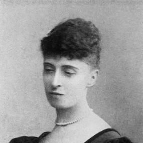 Black and white photograph of Alice Meynell, shown from the shoulders up. She is wearing a black satin dress with puffed sleeves and a sun-shaped pin in the top left corner of the bodice. She has a strand of pearls around her neck and short, dark curly hair.