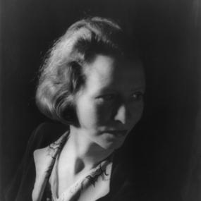 Black and white, shoulder-length photograph of Edna St Vincent Millay by Carl Van Vechten, 1933. She is wearing a black jacket with white collar, and a shirt and necklace. Her hair is cut to jaw-length and her head turned to look down.