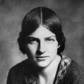 Black-and-white portrait of Naomi Mitchison. Her hair is pinned at the sides of her head in two buns and she is wearing a fuzzy sweater with
            two pom poms at the collar.