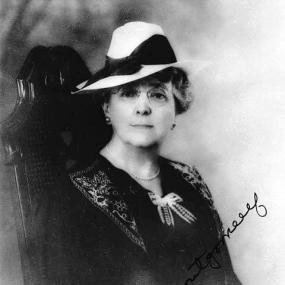 Black and white photograph of L. M. Montgomery, seated in a high-backed chair. Her hair is short, she has round, wire-rimmed glasses. and she looks straight at the camera. She is wearing a black dress with white detail on the collar and a white bow at the neck, a pearl necklace, and a white hat with a wide black riband. Her name is signed across the photo.