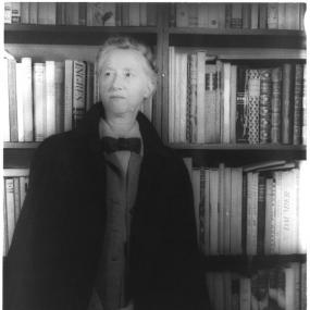 Black and white photograph of Marianne Moore, standing against a bookshelf. Her grey hair is in a plait round her head; she wears a large dark jacket over a grey suit, collared shirt, and bow-tie.