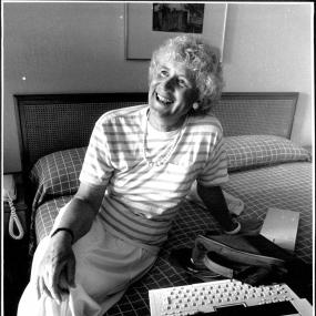 Black-and-white photo of Jan Morris seated on what looks like a hotel bed, smiling. 