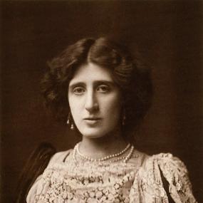 Sepia-toned photograph of Lady Ottoline Morrell, shown from the shoulders up. She is wearing a dark velvet dress with a layer of lace at the top and arms made of semi-sheer ribbed material. Her hair is jaw length, dark, and curly, and she is wearing pearl earrings and necklaces.