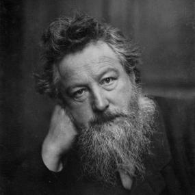 Black and white photograph of William Morris by Frederick Hollyer, c. 1887. Morris, with slanted head, tousled hair, and a beard, looks intently straight forward with his cheek resting on his hand.