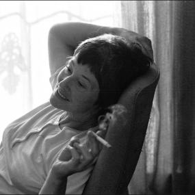 Black-and-white candid photo of Emily Mortimer seated on a chair in front of close blinds with the sun streaming through. She is holding a lit
            cigarette in her right hand and her expression looks serene. 