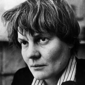 Black-and-white photo of Iris Murdoch in May 1963. Looking intently, off-camera, she is wearing a black blazer with a striped shirt underneath
            and has short hair.