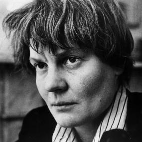 Black-and-white photo of Iris Murdoch in May 1963. Looking intently, off-camera, she is wearing a black blazer with a striped shirt underneath
            and has short hair.