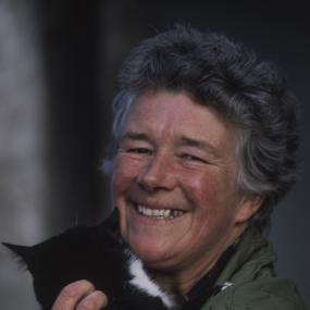 Colour photo of Dervla Murphy smiling and holding a black and white cat that has its face turned away from the camera, October 1990. 