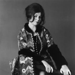 Black-and-white photo of Edna O'brien, December 1970. Seated, she is wearing an eclectic coat with different textual patterns and black fur
            sleeves and collar. Her hair is back in a relaxed bouffant updo.