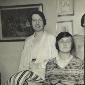 Black and white photograph of Kate O&apos;Brien, sitting in an upholstered chair, with her sister Nancy O&apos;Brien perched on its arm beside her. Both look straight at the camera. Kate is wearing a long, dark skirt and a V-necked sweater with a pattern of horizontal stripes. She has short, dark, straight hair and is holding a paper in her lap.