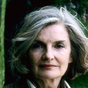 Photo of Julia O'Faolain in Malo, France, 27 May 1986. She is wearing a plaid blazer and a white button-up with a brown silk neck scarf. There
            is greenery behind her. 